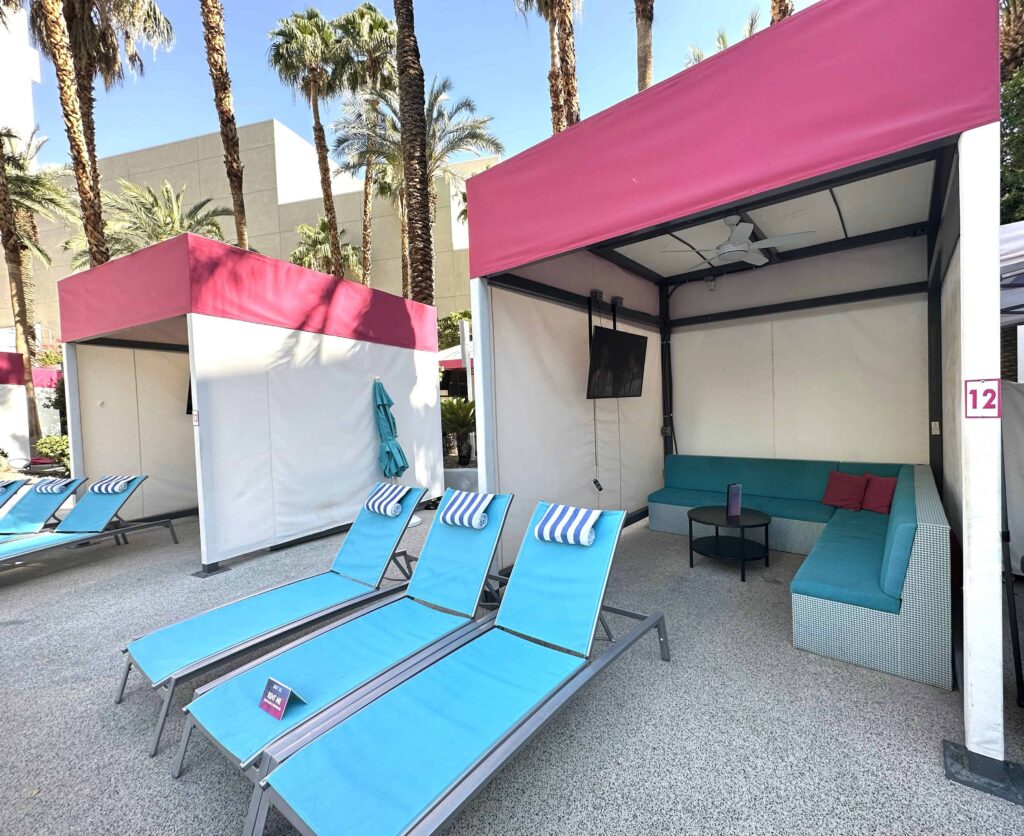 Looking inside a cabana you can see a mounted TV, a couch and a coffee table.   In front are three blue loungers.