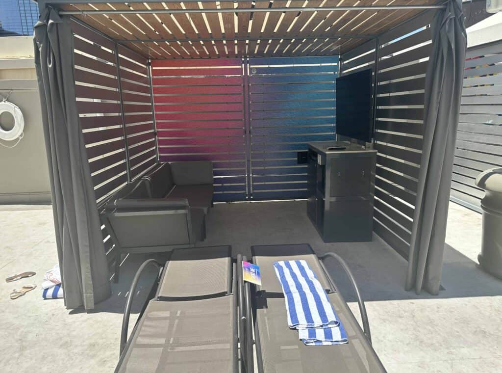 Looking into a cabana you can see a couch and a mounted TV.  There are 2 loungers in front of the cabana.