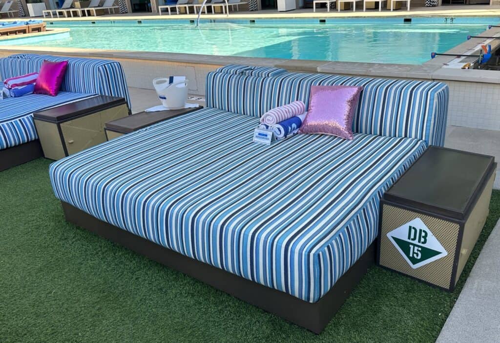 A multicolored striped daybed set on green artificial turf.  A pink pillow sits on the mattress and one of the pools can be seen in the background.