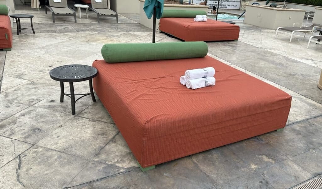 An orange cushioned daybed has rolled towels and a body pillow on it.  A side table can also be seen next to the daybed.